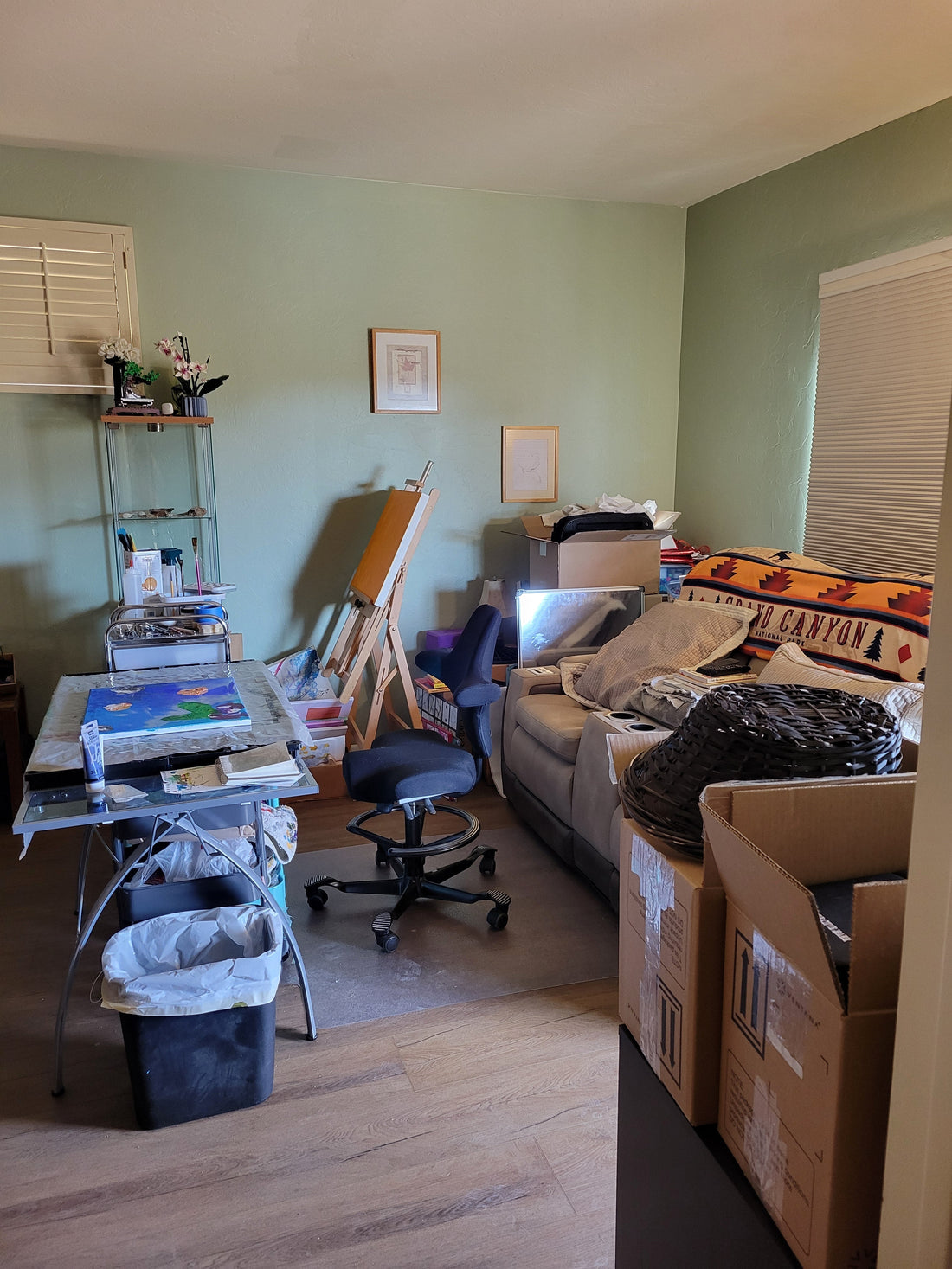 Corner of a large master bedroom. A comfortable and adjustable office chair is flanked by a studio art table and a reclining sofa. Boxes are in the fore and backgrounds and an easel is propped against the far wall.