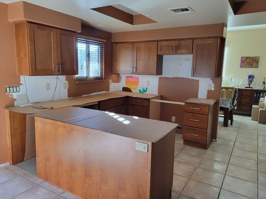 Kitchen in a semi-complete state. There are cabinets but no countertops (we'll discount the semi-functional cardboard flats shown), backsplash, sink, or appliances. In the background, part of the dining room can be seen with a clutter of boxes.