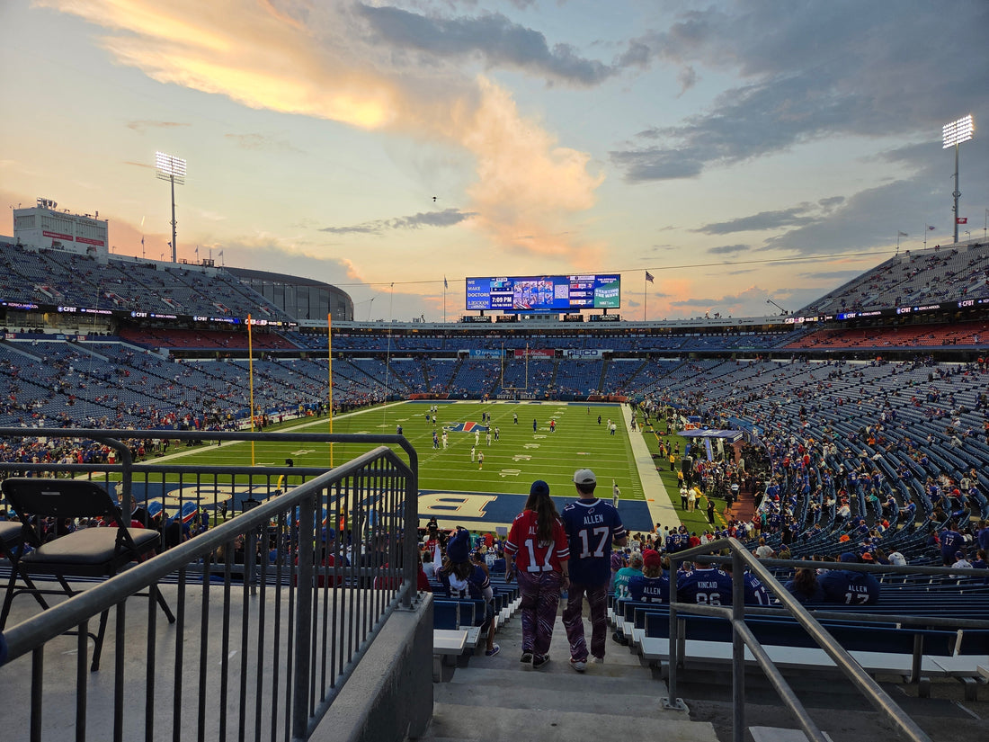 Looking down on the Bills football field from the entrance to an end zone seating section. Two people wearing Josh Allen jerseys (one in red, one in blue) walk down the steps. The sky is a beautiful sunset.