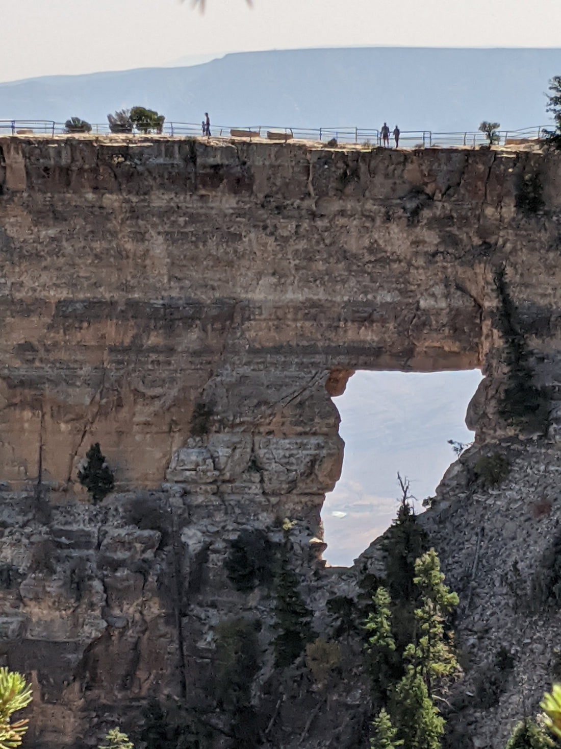 Banded orange, gray, and brown rock with a natural, small arch. Two very small figures stand on the arch's bridge, leaning on railing. Through the arch, the Colorado River glints.