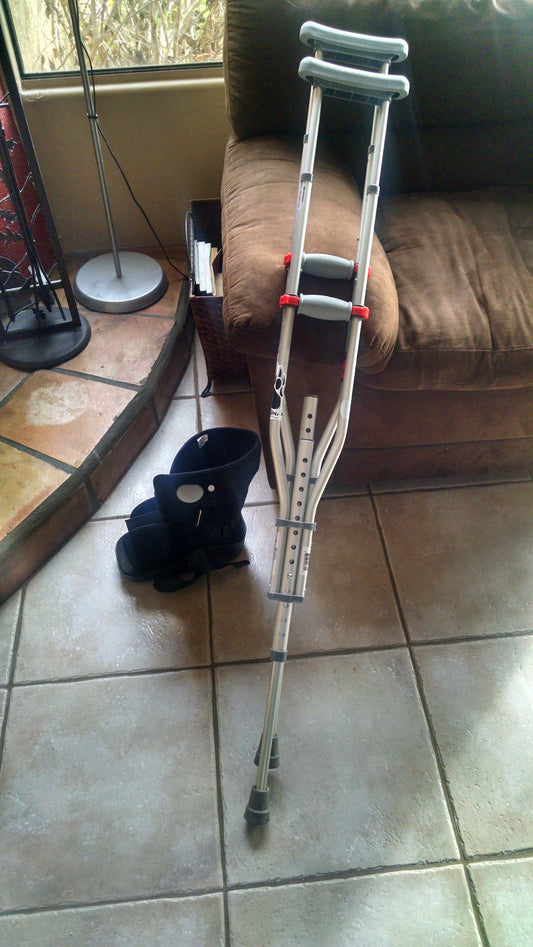 A pair of silver and gray crutches rest against a brown cushiony chair. A black medical walking boot sits on the pinky-beige tile next to the crutches.