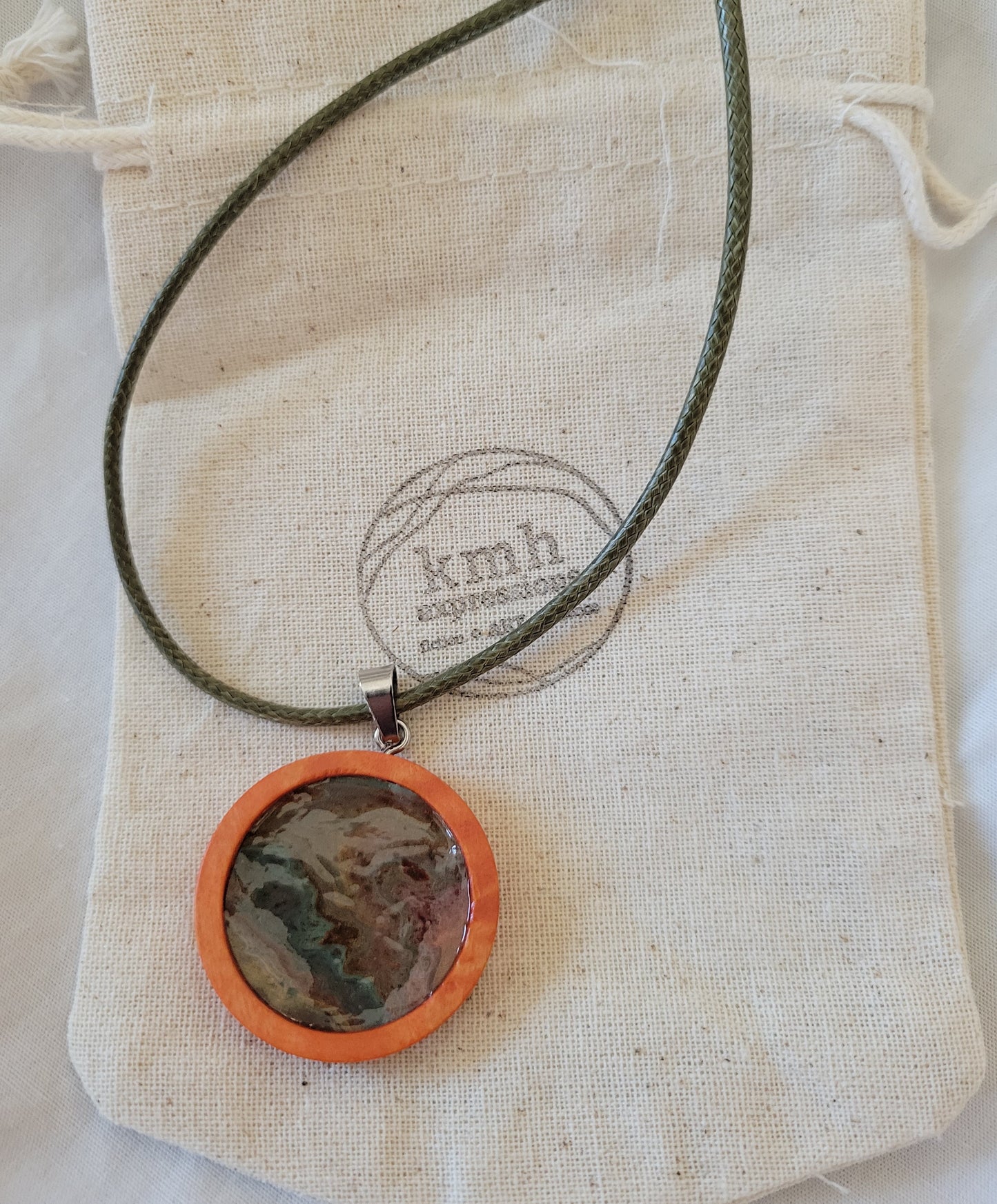 The pendant with a moss green imitation leather cord resting on a canvas bag stamped with the KMH Expressions logo.