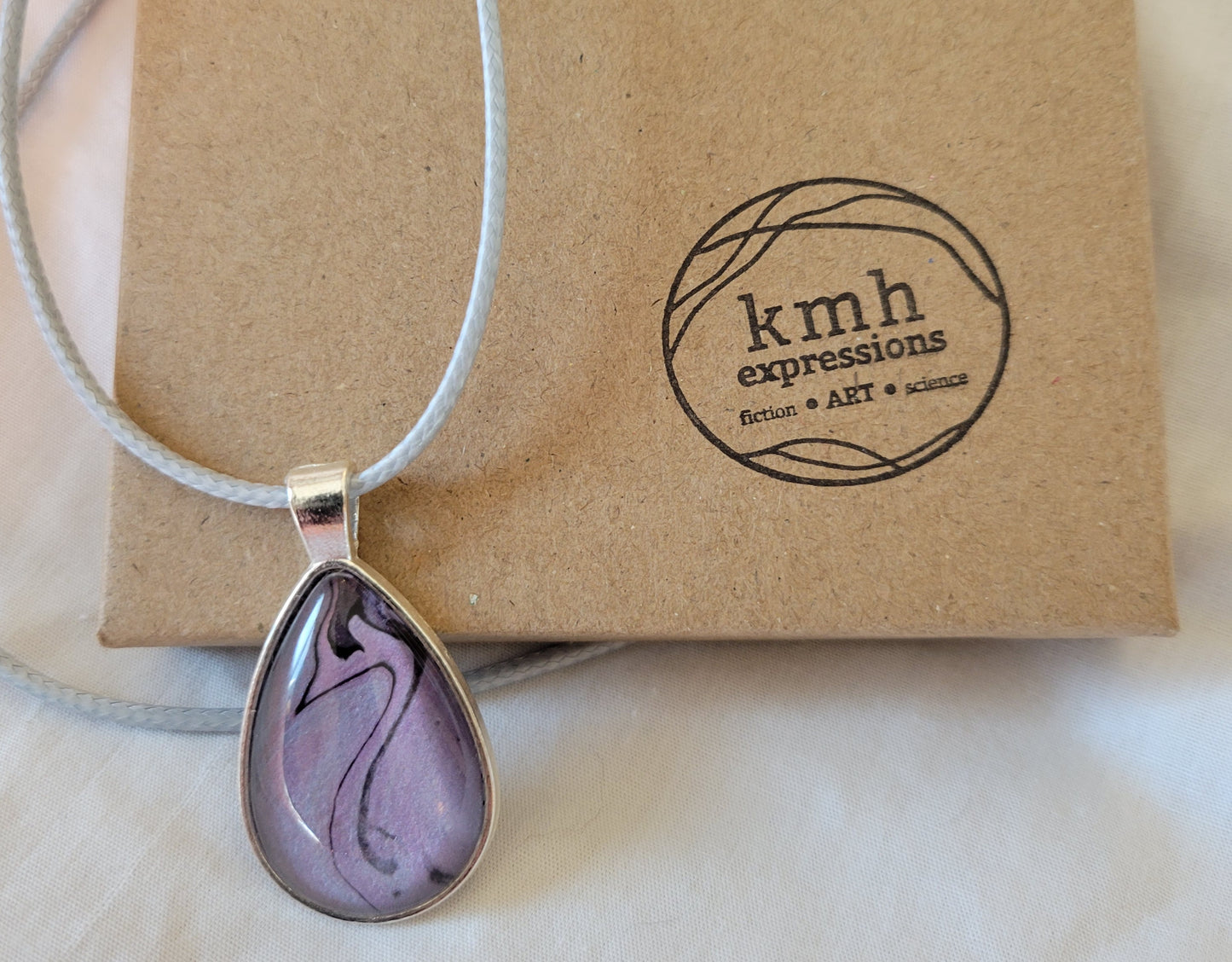 Pendant with a light blue imitation leather cord (that pulls out some hints of blue in the pendant) resting against a cardboard jewelry box stamped with the kmh expressions logo.