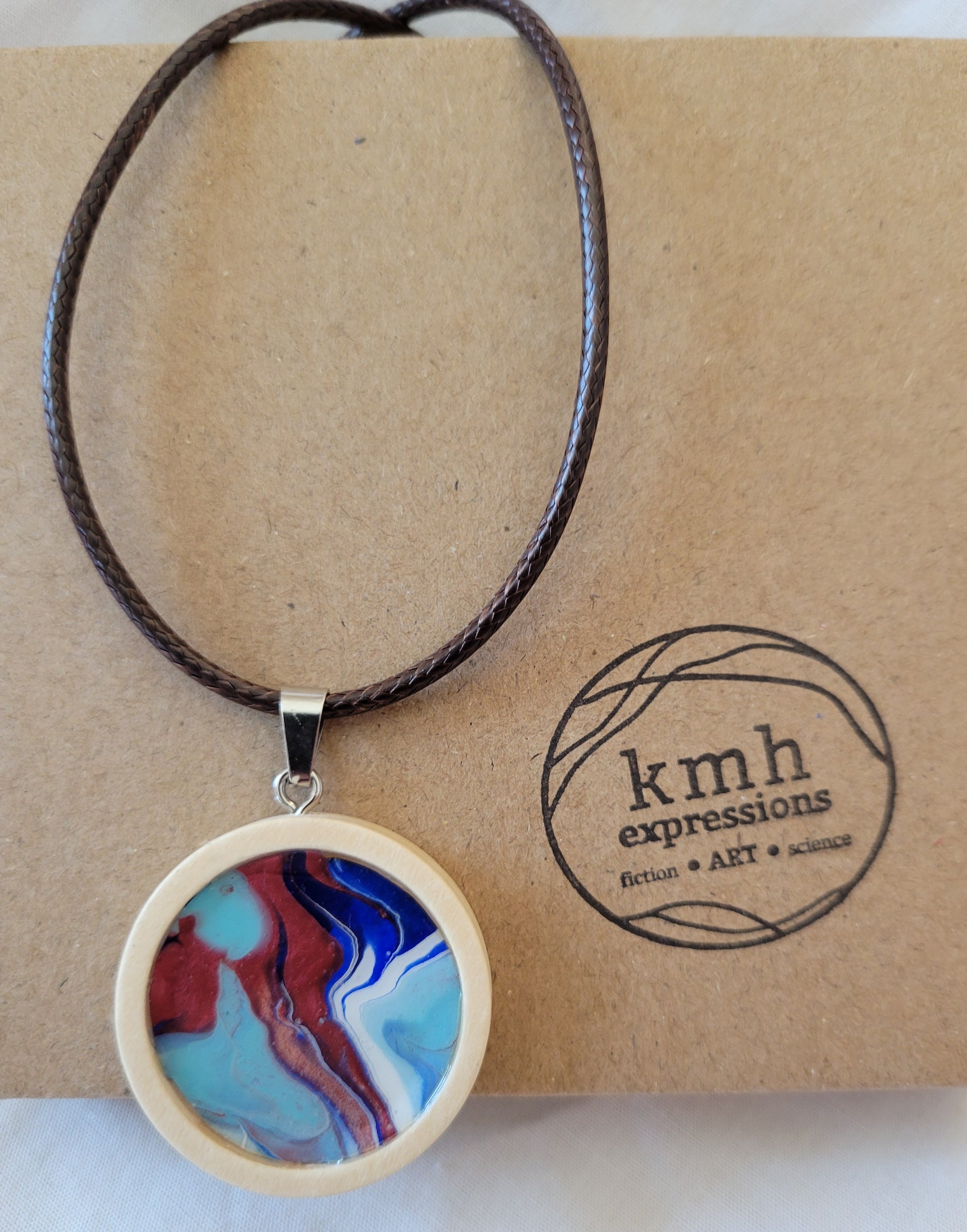 Pendant with a brown imitation leather cord resting against a cardboard jewelry box stamped with the kmh expressions logo.