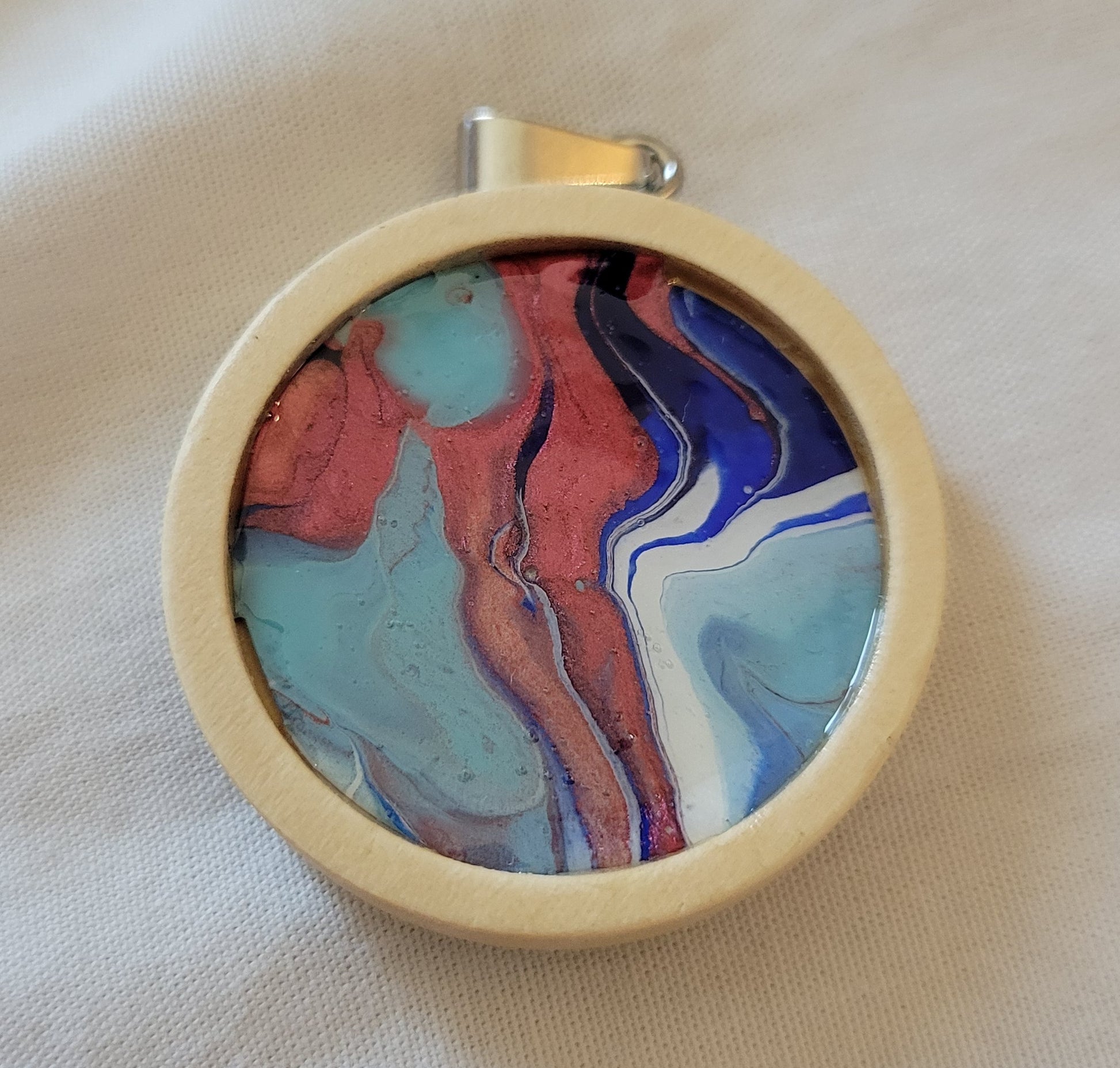 Circular pendant with a simple off-white wood setting and silver hardware. Pendant art is a blend of coppery-red, light blue turquoise, dark blue, and white in a warped banded pattern.