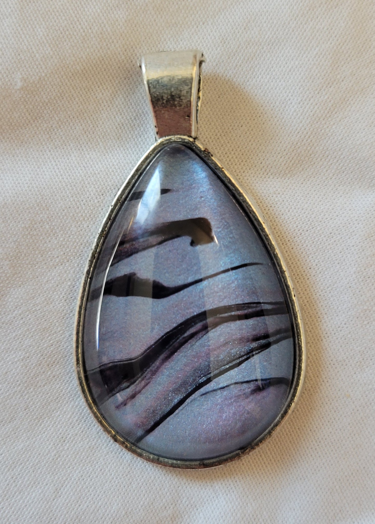 A simple stainless steel teardrop pendant. The overall light shiny blue color is softly streaked through with light pink and violet. There are stark black lines threaded through the colors that evoke rolling hills with a jagged ancient ruin on top of them.