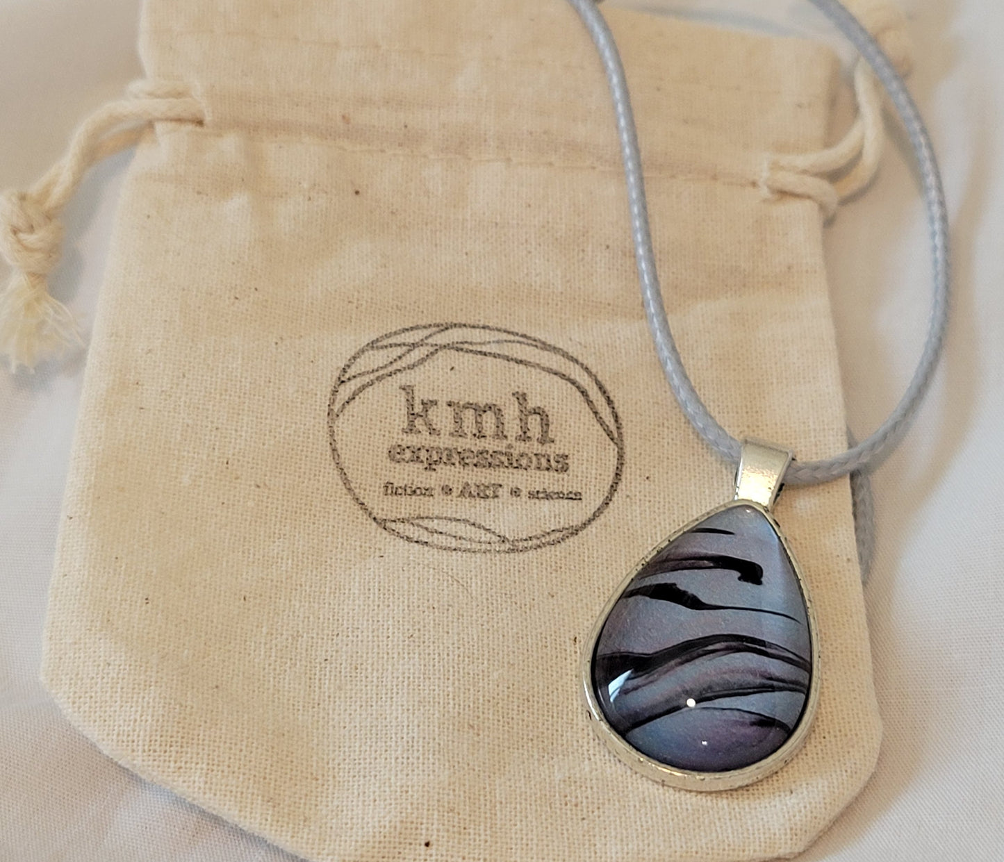 Simple teardrop stainless steel pendant with a light blue imitation leather cord resting on a muslin bag stamped with the kmh expressions logo. The pendant is largely a light blue with hints of violet and faint pink and streaked through with stark black.