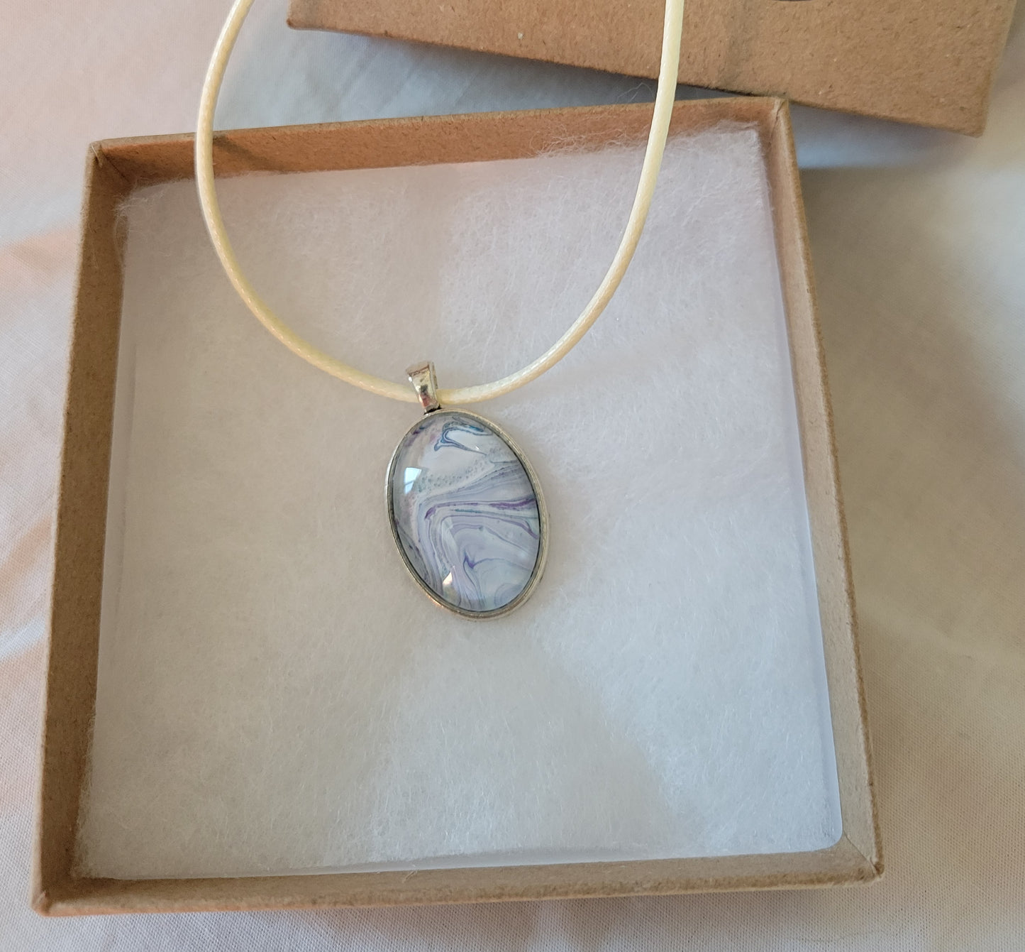 A simple oval pendant with a cream colored imitation leather cord rests on gauze in a brown cardboard jewelry box.