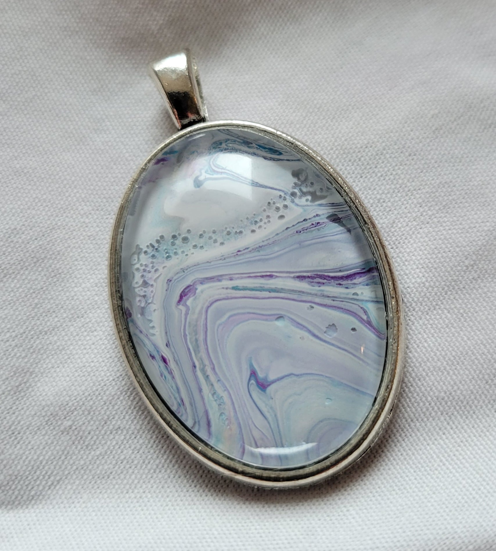 A simple stainless steel oval pendant. White, green-blue, and purple are layered over each other and banded against each other in a half-swirl, the dominant white becoming cloudy with the other colors.