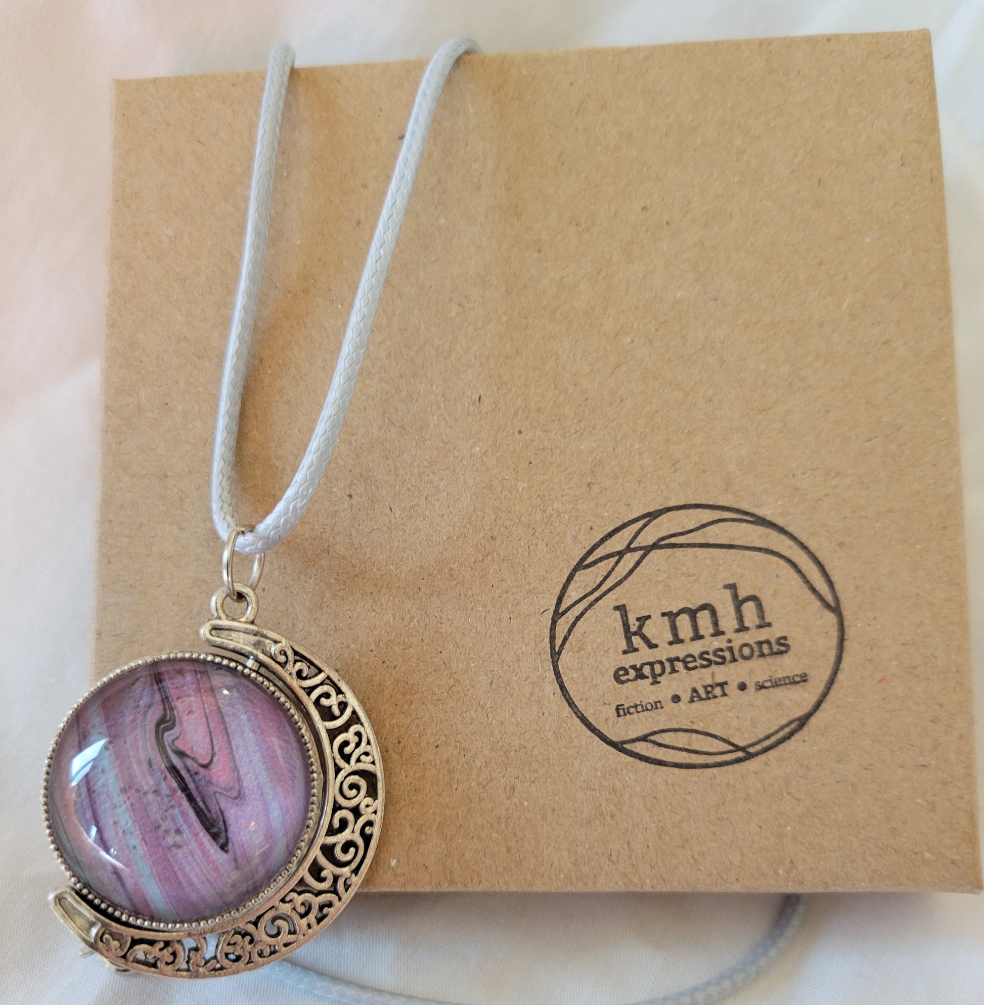 The dual-sided swivel circle pendant strung artfully by its light blue imitation leather cord against a cardboard jewelry box with the logo for kmh expressions. The side of the pendant shown is mostly light purple and pink with threads of blue and black visible.