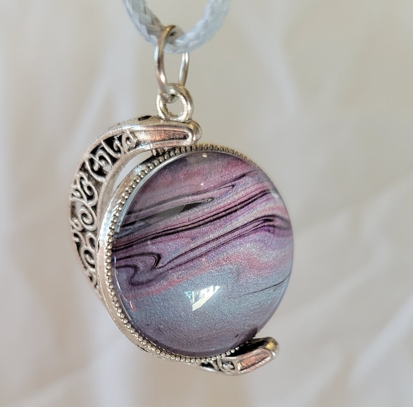 Clouds of gas swirl in the atmosphere of this tilted spinning planet. Stainless steel filigree arcs around the swivel points of this pendant. On this side of the pendant, a light blue dominates the lower portion while pinks, purples, and black weave and swirl through and around each other at the top.