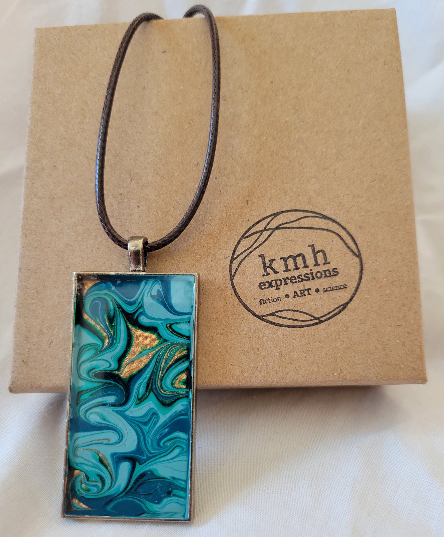 A simple bronze alloy pendant setting with patterned greens and blues and gold dangling from a brown imitation leather cord draped on a brown cardboard jewelry box stamped with the kmh expressions logo.